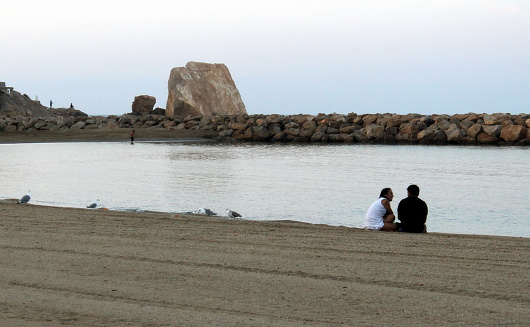 Águilas, Murcia, Spain; On August 12, 2018: Two people sitting on sand and talking very close to each other next to the seashore with a stone breakwater and a big rock in the background