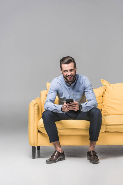 istock cheerful man using smartphone and sitting on yellow sofa on grey 1046941064