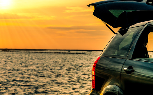 Black compact SUV car with sport and modern design parked on concrete road by the sea at sunset. Environmentally friendly technology. Road trip travel on vacation at the beach and open car truck