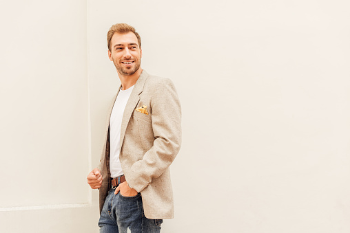 smiling handsome man in jacket and jeans standing near beige wall in city