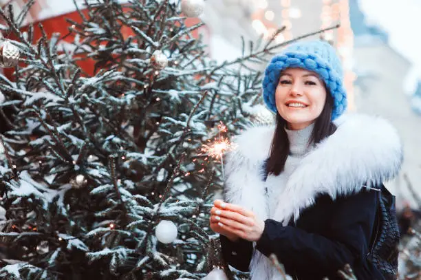 christmas portrait of happy woman with burning firelight walking outdoor in snowy winter city in fashion coat with white fur and oversize chunk knit hat