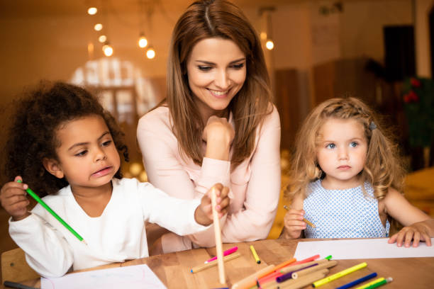 istock cheerful educator sitting near multicultural kids drawing in kindergarten 1047023906