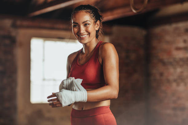 Woman getting ready for boxing practice Woman boxer wearing strap on wrist for boxing practice. Fitness female getting ready for boxing practice. Beautiful young woman with muscular body preparing for workout looking away and smiling. kickboxing photos stock pictures, royalty-free photos & images
