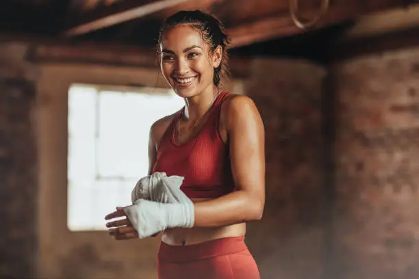 Woman boxer wearing strap on wrist for boxing practice. Fitness female getting ready for boxing practice. Beautiful young woman with muscular body preparing for workout looking away and smiling.