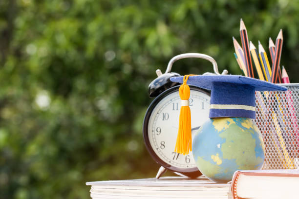 istock Graduate or Education knowledge study abroad concept : Graduation cap on textbook with pencil box and retro clock on desk, blur green light background. Back to School 1047048396