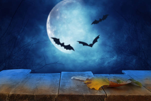 istock Halloween holiday concept. Empty rustic table in front of scary and misty night sky with black bats and full moon background. Ready for product display montage. 1047053856