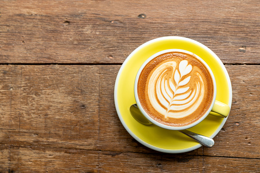 Top view of hot cappuccino coffee in a yellow cup with latte art and saucer on the wooden table background.