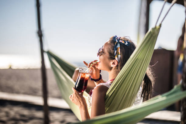 szczęśliwa kobieta relaksująca się w hamaku na plaży i ciesząc się letnim napojem. - juice drinking women drink zdjęcia i obrazy z banku zdjęć