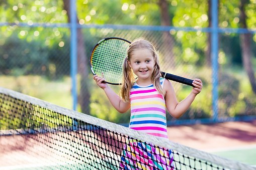 Child playing tennis on outdoor court. Little girl with tennis racket and ball in sport club. Active exercise for kids. Summer activities for children. Training for young kid. Child learning to play.
