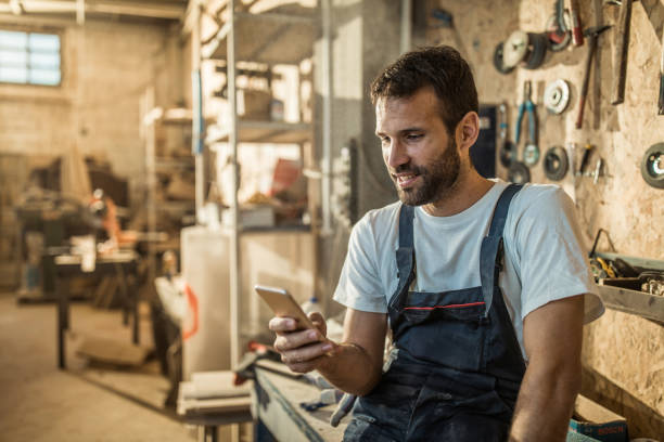 Happy manual worker using mobile phone in a workshop. Manual worker surfing the Internet on a cell phone at workshop. repairperson occupation stock pictures, royalty-free photos & images