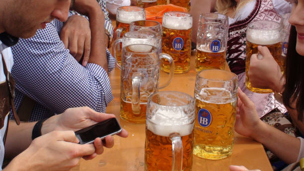 Oktoberfest Celebration In Munich Germany And People Drinking Beer Scene Of Advertisement Sign, Lot Of People Wearing Bavarian Traditional Oktoberfest Costumes Sitting Down In A Beer Tent, Looking Around, Reading Text Message With Smartphone, Taking Picture, Talking To One Another, Laughing, Holding Giant Beer Mug And Drinking Beer During Annual Oktoberfest In Munich Germany Europe large german beer steins stock pictures, royalty-free photos & images