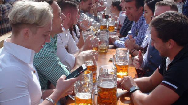 Oktoberfest In Munich Germany Plus People Sitting Down And Having A Drink Scene Of Advertisement Sign, Lot Of People Wearing Bavarian Traditional Oktoberfest Costumes Sitting Down In A Beer Tent, Looking Around, Reading Text Message With Smartphone, Taking Picture, Talking To One Another, Laughing, Holding Giant Beer Mug And Drinking Beer During Annual Oktoberfest In Munich Germany Europe large german beer steins stock pictures, royalty-free photos & images