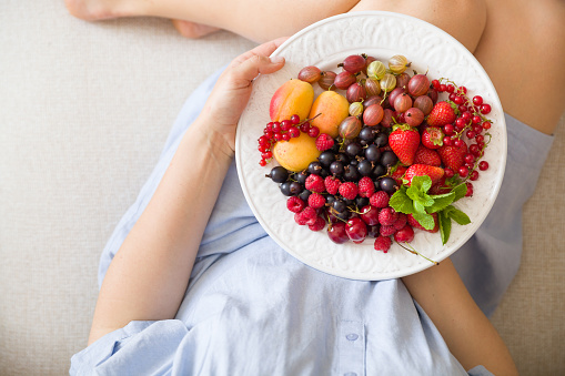 Top view on woman sitting cozy on the couch and enjoying delicious summer fruit and berries. Girl holding white plate with apricots, raspberries, strawberries, blackcurrant, blueberries on her knees