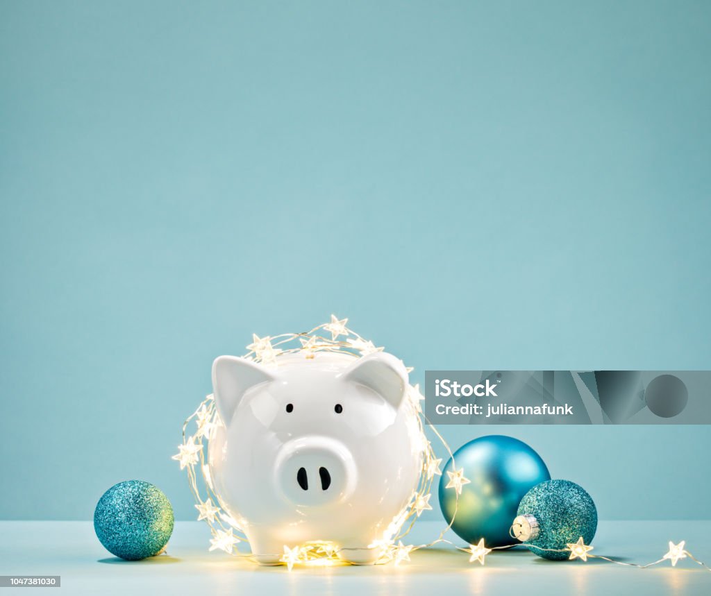 Piggy bank wrapped in a string of Christmas lights White Piggy bank wrapped in a string of Christmas lights over a blue background. Saving concept. Christmas Stock Photo