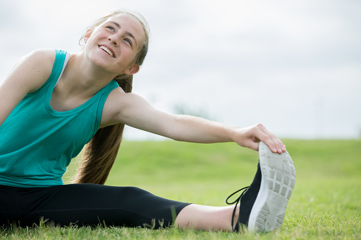 Beautiful Caucasian teenage girl smiles and looks toward the sky as she stretches her muscles before running outside in green grass.