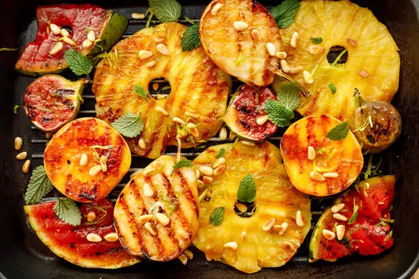 Photo of Grilled fruits; pineapples, peaches, figs, pears and watermelon with pine nuts, fresh herbs and honey on the grill plate, top view.