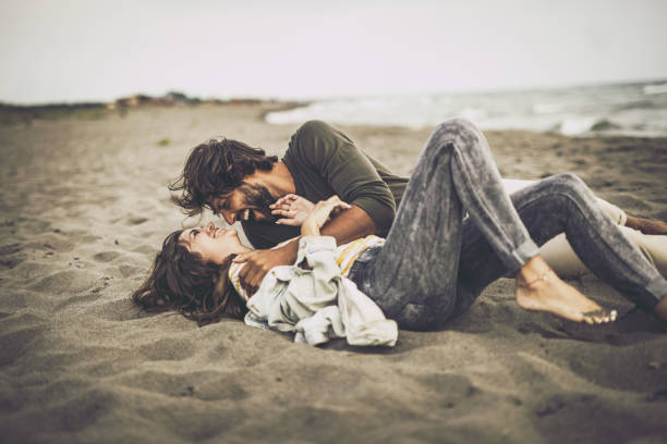 Young cheerful couple having fun while lying in sand during autumn day at the beach.