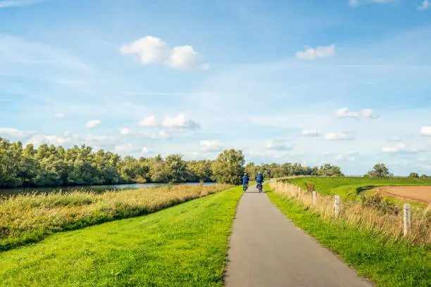 Older man and woman cycle on a curved cycle path on a dike on the edge of the Dutch National Park De Biesbosch. It is a sunny day with white clouds on the blue sky in the beginning of the fall season.