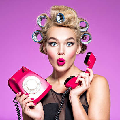 young woman with wonder face holds retro phone. Girl with blue curlers talking by red telephone. Young woman with blue curlers calling by retro phone.