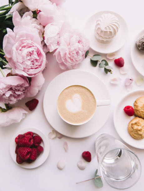 istock Delicious morning breakfast table essentials: fresh cappuccino coffee, marshmallow, cookies, chocolate, red raspberries, glass of water and tender pink blossoming peony bouquet on the white background 1047503196