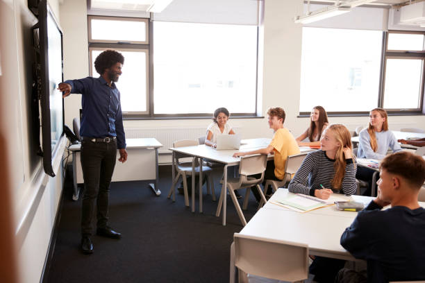 istock Male High School Teacher Standing Next To Interactive Whiteboard And Teaching Lesson 1047530118