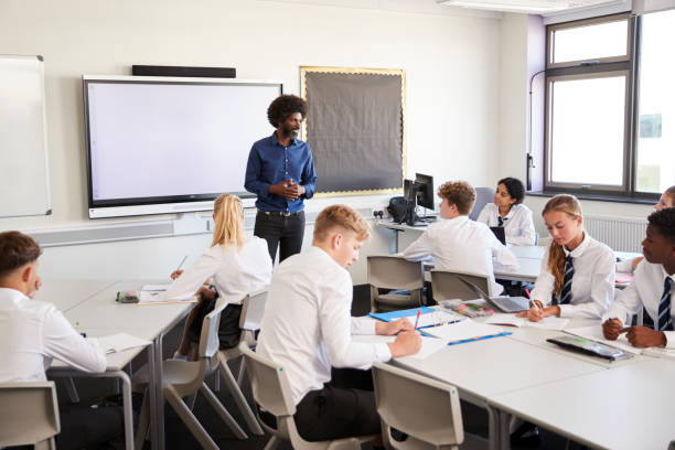 istock Male High School Teacher Standing Next To Interactive Whiteboard And Teaching Lesson To Pupils Wearing Uniform 1047532630
