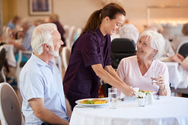 Senior Couple Being Served With Meal By Carer In Dining Room Of Retirement Home Senior Couple Being Served With Meal By Carer In Dining Room Of Retirement Home retirement community stock pictures, royalty-free photos & images