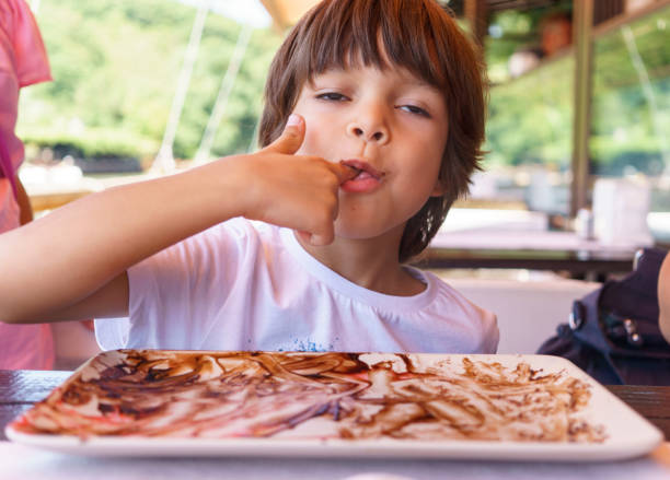 A playful young boy with a messy face enjoys a chocolate treat, surrounded by empty plates and cups. His mischievous grin and chocolate-covered hands show he's had a fun time eating.