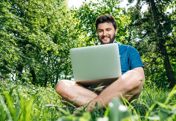 istock Young modern businessman freelancer working on computer and sitting on grass. Male student using notebook outdoors. Tourist working on laptop 1047570570