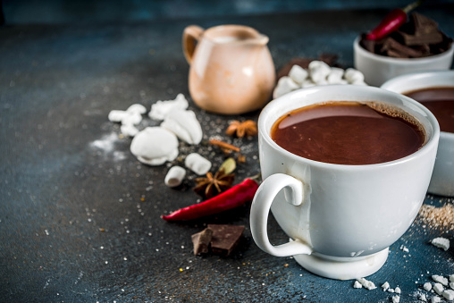 Hot chocolate cups, broken pieces of chocolate, sugar, marshmallow, red chili peppers and spices. Dark blue concrete background top view space