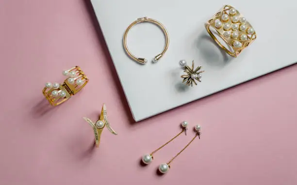 Photo of golden bracelets and earrings with pearls on pink and white background