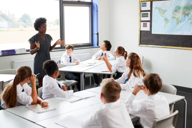 istock Female High School Tutor Standing By Tables With Students Wearing Uniform Teaching Lesson 1047617460