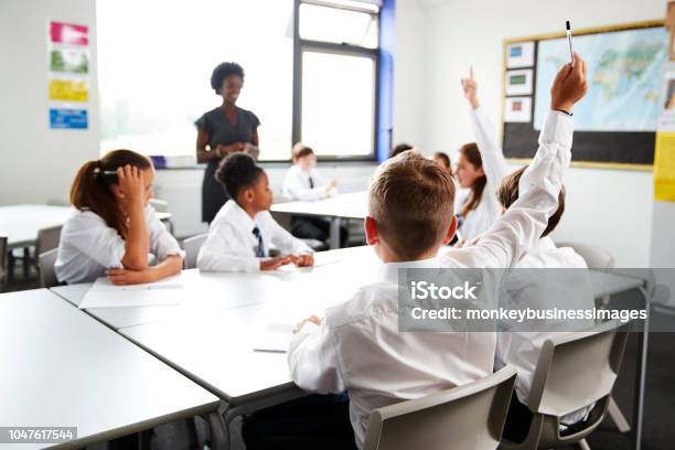 High School Students Wearing Uniform Raising Hands To Answer Question Set By Teacher In Classroom Stock Photo - Download Image Now