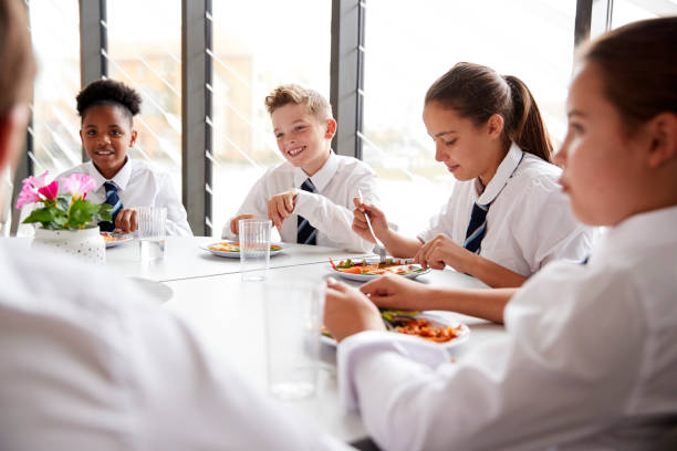istock Group Of High School Students Wearing Uniform Sitting Around Table And Eating Lunch In Cafeteria 1047619904