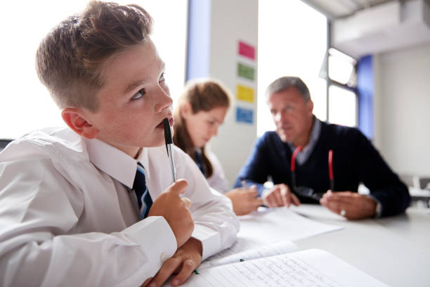 istock Concentrating Male High School Student Wearing Uniform Working At Table With Teacher Talking To Pupils In Background 1047620322