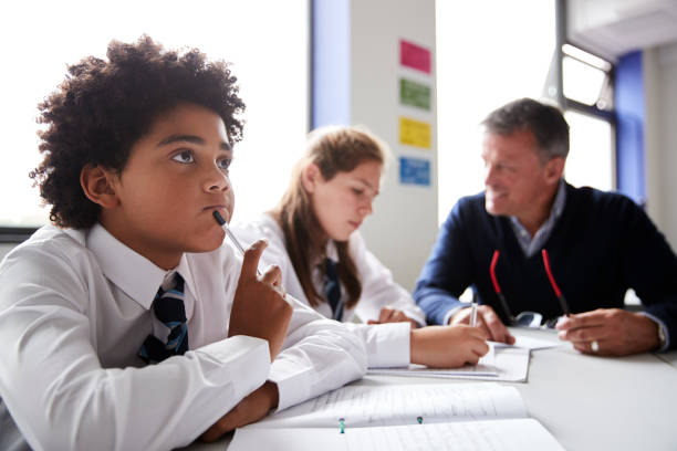 istock Concentrating Male High School Student Wearing Uniform Working At Table With Teacher Talking To Pupils In Background 1047620332