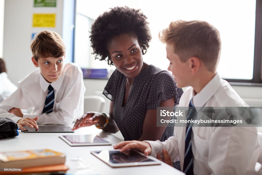 Professor de liceu feminino sentado à mesa com os alunos usando uniforme usando comprimidos Digital na lição - Foto de stock de Professor royalty-free