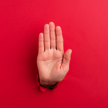 Human hand reaching through torn red paper sheet showing 