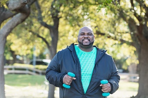 A mature African-American man in his 40s with shaved head, mustache and beard, exercising in the park with hand weights, smiling at the camera. He has a large build.