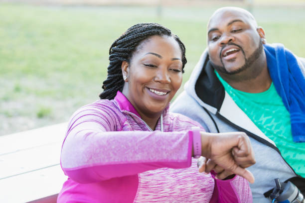 African-American couple, checking fitness tracker Close-up of an African-American couple at the park exercising. The mid adult woman, in her 30s, is showing her partner, a mature man in his 40s, her fitness tracker. The focus is on her smiling face. body positive couple stock pictures, royalty-free photos & images