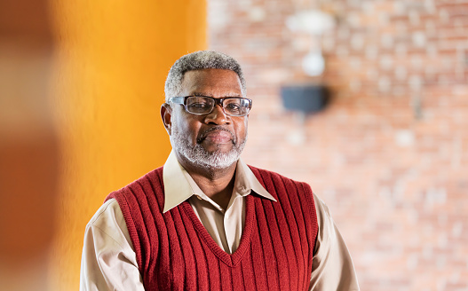 A senior African-American man in his 60s wearing eyeglasses, a button down shirt and sweater vest, standing indoors, looking at the camera with a serious, confident expression.