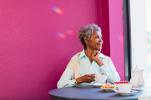 A senior African-American woman in her 70s sitting at a table in a coffee shop, having coffee and pastries, smiling as she looks out the window.