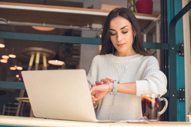 jovem fêmea freelancer em fones de ouvido verificando o relógio de pulso na mesa com o laptop e vinho quente no café - mulled wine audio - fotografias e filmes do acervo