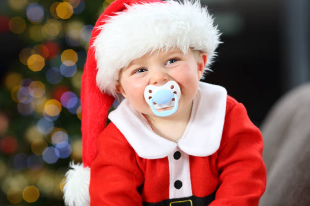 Baby wearing santa disguise with a pacifier in the mouth looking at camera in christmas on a couch in the living room at home