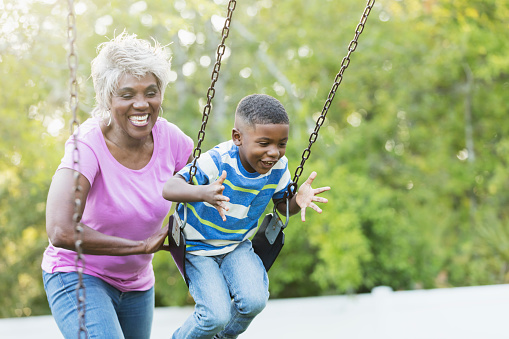 A senior African-American woman in her 60s with her 4 year old grandson at the park. She is pushing him on a swing on the playground, and he is having fun.