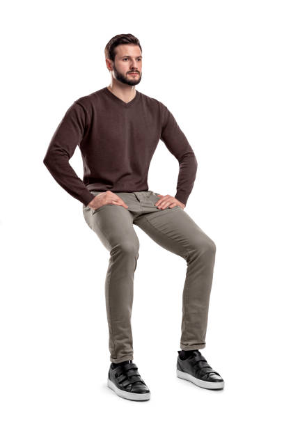 An isolated bearded man in casual wear sits on a white background with hands on his thighs. An isolated bearded man in casual wear sits on a white background with hands on his thighs. Sitting and waiting. Meditating. Neutral expression. sitting stock pictures, royalty-free photos & images