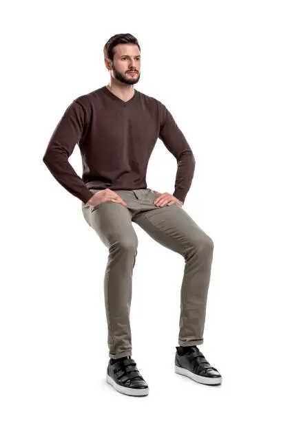 An isolated bearded man in casual wear sits on a white background with hands on his thighs. Sitting and waiting. Meditating. Neutral expression.