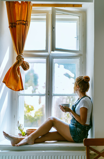 Young beautiful woman is looking through the window and drinking coffee in the morning