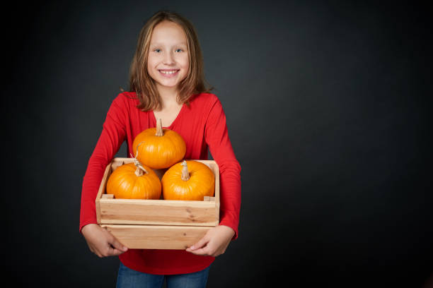 Smiling girl holding a box with orange pumpkins, over dark background