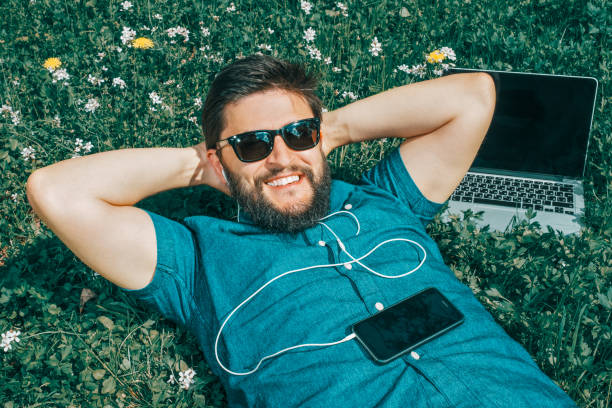 istock Tired freelancer lying on grass near his laptop computer. Handsome man in sunglasses resting with his hands behind the head 1047710828